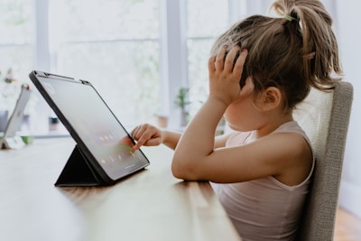A young child with their hair in a ponytail sits at a table, interacting with a tablet device. The child rests one hand against their face while using their other hand to touch the screen. Natural light filters through large windows in the background, creating a soft and calm environment.