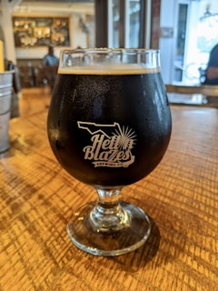 A glass of dark beer sits on a wooden table in a cozy indoor setting. The glass features the logo of 'Hell 'n Blazes Brewing Co.' in white lettering. The background shows an out-of-focus bar area with some people and decor visible.