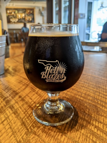A glass of dark beer sits on a wooden table in a cozy indoor setting. The glass features the logo of 'Hell 'n Blazes Brewing Co.' in white lettering. The background shows an out-of-focus bar area with some people and decor visible.
