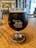 A glass of dark beer sits on a wooden table in a cozy indoor setting. The glass features the logo of 'Hell 'n Blazes Brewing Co.' in white lettering. The background shows an out-of-focus bar area with some people and decor visible.