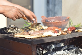 Hands are seen grilling a variety of meats and vegetables over a barbecue with visible charcoals and smoke. A clay dish and some green herbs are also present in the background.