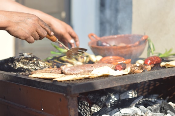 Hands are seen grilling a variety of meats and vegetables over a barbecue with visible charcoals and smoke. A clay dish and some green herbs are also present in the background.