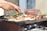 Hands are seen grilling a variety of meats and vegetables over a barbecue with visible charcoals and smoke. A clay dish and some green herbs are also present in the background.