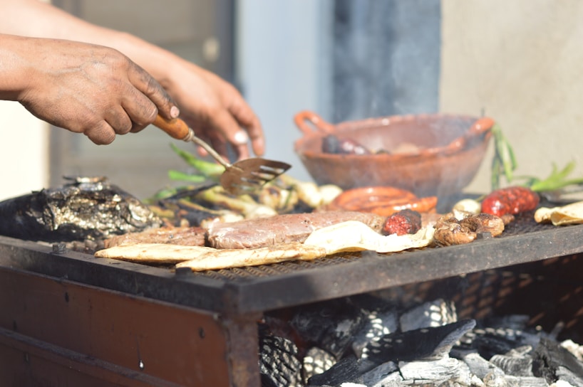 Hands are seen grilling a variety of meats and vegetables over a barbecue with visible charcoals and smoke. A clay dish and some green herbs are also present in the background.