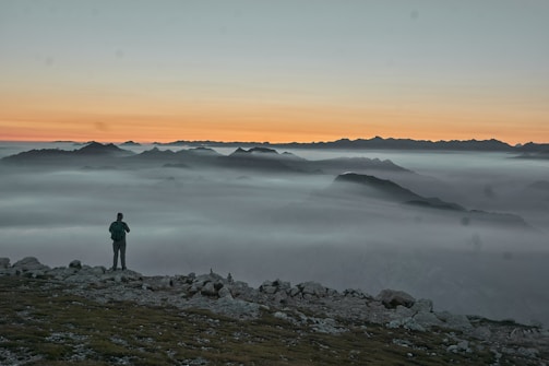 A hiker standing on a rocky peak overlooking a vast, misty mountain range at sunrise.