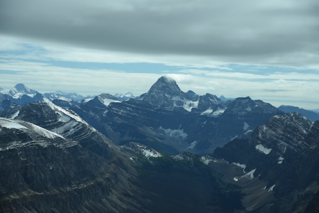 A majestic mountain range with snow-capped peaks under an overcast sky. The central peak stands prominently above the others, creating a dramatic and awe-inspiring landscape. The surrounding mountains feature steep cliffs and ridges with patches of snow.