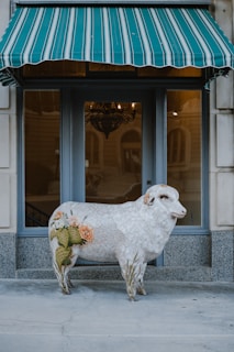 A decorative sculpture of a sheep with intricate patterns and designs adorns its body, including cacti and roses. It is positioned in front of a window with a green and white striped awning above. The background includes a building facade and a chandelier inside the window.