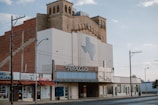 An old theater building with an art deco style facade stands on a quiet street. A large sign featuring the word 'Texas' and a map outline of the state decorates the front. The building has a brick structure with decorative windows and vintage signage. Nearby are small shops with signs, including a thrift shop.