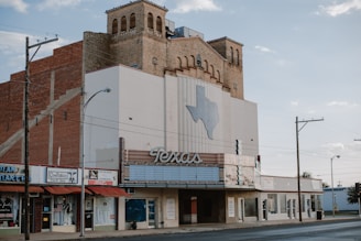 An old theater building with an art deco style facade stands on a quiet street. A large sign featuring the word 'Texas' and a map outline of the state decorates the front. The building has a brick structure with decorative windows and vintage signage. Nearby are small shops with signs, including a thrift shop.