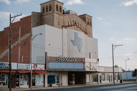 An old theater building with an art deco style facade stands on a quiet street. A large sign featuring the word 'Texas' and a map outline of the state decorates the front. The building has a brick structure with decorative windows and vintage signage. Nearby are small shops with signs, including a thrift shop.