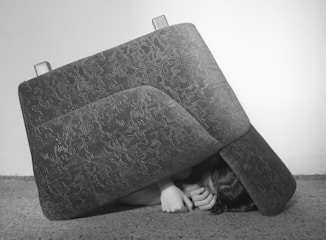 A person is lying on the floor partially covered by an upturned couch cushion with an abstract pattern. The person's hands and part of the face are visible, appearing as if they are sheltering or hiding. The background is a plain wall, and the floor is carpeted.