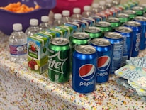 A colorful party setup with a table covered in a confetti-patterned tablecloth. The table is lined with beverages, including cans of Pepsi and Sprite, cartons of apple juice, and small water bottles. A bowl of orange snacks, potentially cheese puffs, is in the background, and a stack of napkins with 'Party Time!' printed on them is in the foreground.
