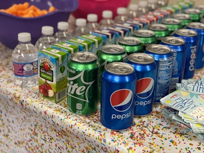A colorful party setup with a table covered in a confetti-patterned tablecloth. The table is lined with beverages, including cans of Pepsi and Sprite, cartons of apple juice, and small water bottles. A bowl of orange snacks, potentially cheese puffs, is in the background, and a stack of napkins with 'Party Time!' printed on them is in the foreground.