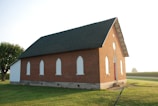 A small, traditional brick church with white-framed arched windows and a steep green roof. It sits on a well-maintained grassy lawn under a clear sky. The building has a quaint, historical charm, with a single red door at the front.