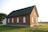 A small, traditional brick church with white-framed arched windows and a steep green roof. It sits on a well-maintained grassy lawn under a clear sky. The building has a quaint, historical charm, with a single red door at the front.