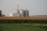 A peaceful rural landscape featuring large silos and a barn amidst expansive fields of corn and soybeans. The structures stand tall against a backdrop of distant trees and a hazy sky, suggesting early morning or late afternoon light.