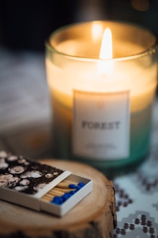 A lit candle in a glass holder labeled 'Forest' emits a warm glow, placed on a wooden surface. In the foreground, a matchbox with a floral design contains blue-tipped matches.