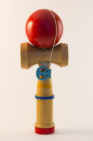 A wooden toy featuring a red ball connected by a string to a handle. The handle has colorful bands in blue, yellow, and red, and includes a small decorative blue piece. The toy is positioned vertically against a plain background.