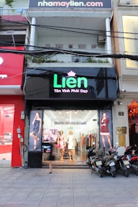 A storefront named 'Lien' with mannequins dressed in fashionable clothing placed in the window. The exterior displays a black sign with green and pink lettering. Several large posters of models in dresses flank the entrance. Scooters are parked on the sidewalk in front of the store.