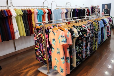 Various colorful dresses are neatly arranged on display racks within a brightly lit clothing store. The garments feature a range of patterns, including floral and abstract designs, hanging on wooden hangers. The floor is polished wood, and the room has a clean, organized appearance.