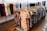 Various colorful dresses are neatly arranged on display racks within a brightly lit clothing store. The garments feature a range of patterns, including floral and abstract designs, hanging on wooden hangers. The floor is polished wood, and the room has a clean, organized appearance.