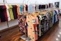 Various colorful dresses are neatly arranged on display racks within a brightly lit clothing store. The garments feature a range of patterns, including floral and abstract designs, hanging on wooden hangers. The floor is polished wood, and the room has a clean, organized appearance.