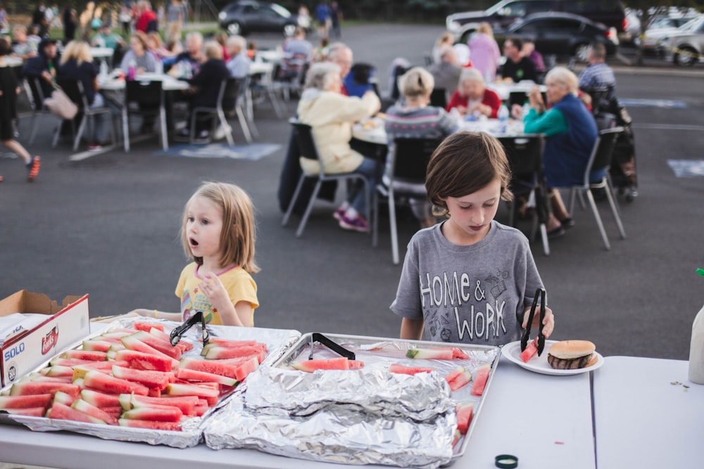 a couple of kids standing next to a table filled with food