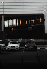 A modern building with sleek lines and illuminated vertical lights on the facade. In front, there are parked cars including a white BMW and a black Volvo. A few people are visible near the entrance, standing by the railing. The scene appears to be in a dimly lit urban setting.