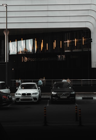 A modern building with sleek lines and illuminated vertical lights on the facade. In front, there are parked cars including a white BMW and a black Volvo. A few people are visible near the entrance, standing by the railing. The scene appears to be in a dimly lit urban setting.