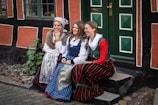 Three women dressed in traditional folk costumes are seated on a stone step in front of a rustic building with vibrant orange and black walls. The women appear to be engaged in a lighthearted conversation, smiling naturally. The door behind them is painted green with a distinct geometric pattern. Plants and cobblestones add to the quaint, old-world charm of the setting.