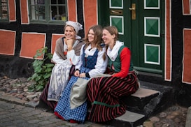Three women dressed in traditional folk costumes are seated on a stone step in front of a rustic building with vibrant orange and black walls. The women appear to be engaged in a lighthearted conversation, smiling naturally. The door behind them is painted green with a distinct geometric pattern. Plants and cobblestones add to the quaint, old-world charm of the setting.