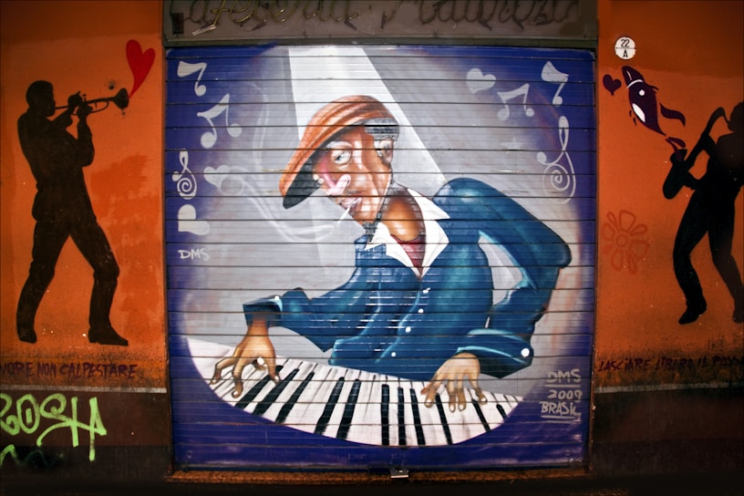 A vivid graffiti mural depicting a musician playing a keyboard. The musician is wearing a hat and appears to be deeply engrossed in the music. Musical notes and hearts are painted around, suggesting a lively musical theme. Silhouettes of a trombone player and a saxophonist frame the central image, emphasizing a jazz motif. The mural has a dynamic composition with vibrant colors.