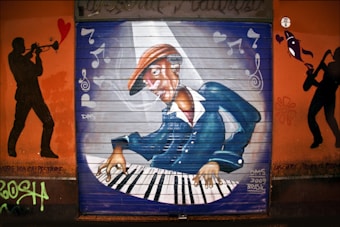 A vivid graffiti mural depicting a musician playing a keyboard. The musician is wearing a hat and appears to be deeply engrossed in the music. Musical notes and hearts are painted around, suggesting a lively musical theme. Silhouettes of a trombone player and a saxophonist frame the central image, emphasizing a jazz motif. The mural has a dynamic composition with vibrant colors.
