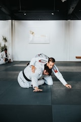 Two people are practicing martial arts on a padded floor. They are wearing white uniforms with black belts, indicating a level of proficiency. One person is on top of the other, controlling the movement. The setting appears to be a martial arts studio with a minimalistic interior, including white walls and a dark ceiling. A plant is visible in the background, adding a touch of greenery to the scene.