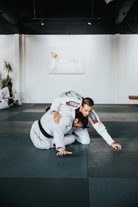 Two people are practicing martial arts on a padded floor. They are wearing white uniforms with black belts, indicating a level of proficiency. One person is on top of the other, controlling the movement. The setting appears to be a martial arts studio with a minimalistic interior, including white walls and a dark ceiling. A plant is visible in the background, adding a touch of greenery to the scene.