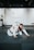 Two people are practicing martial arts on a padded floor. They are wearing white uniforms with black belts, indicating a level of proficiency. One person is on top of the other, controlling the movement. The setting appears to be a martial arts studio with a minimalistic interior, including white walls and a dark ceiling. A plant is visible in the background, adding a touch of greenery to the scene.