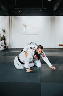 Two people are practicing martial arts on a padded floor. They are wearing white uniforms with black belts, indicating a level of proficiency. One person is on top of the other, controlling the movement. The setting appears to be a martial arts studio with a minimalistic interior, including white walls and a dark ceiling. A plant is visible in the background, adding a touch of greenery to the scene.