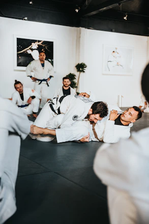 Practitioners wearing white martial arts uniforms are engaged in a grappling session on a mat in a training facility. Other participants and an instructor are watching the exercise. The room includes framed photos on the walls and a potted plant in the corner.