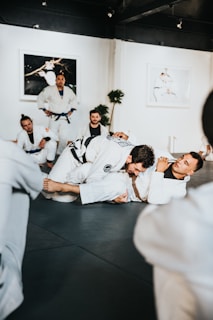 Practitioners wearing white martial arts uniforms are engaged in a grappling session on a mat in a training facility. Other participants and an instructor are watching the exercise. The room includes framed photos on the walls and a potted plant in the corner.