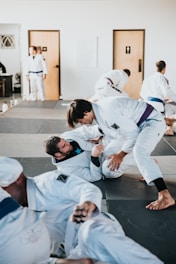 Several individuals are practicing martial arts in a gym setting. They are engaged in a grappling exercise on padded mats, wearing traditional white uniforms with colored belts. The room has doors in the background and appears well-lit.