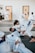Several individuals are practicing martial arts in a gym setting. They are engaged in a grappling exercise on padded mats, wearing traditional white uniforms with colored belts. The room has doors in the background and appears well-lit.