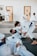 Several individuals are practicing martial arts in a gym setting. They are engaged in a grappling exercise on padded mats, wearing traditional white uniforms with colored belts. The room has doors in the background and appears well-lit.