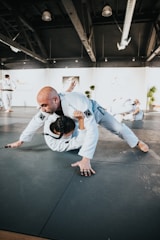 Two martial artists are practicing grappling techniques on a mat in a spacious training facility. The scene takes place under a high ceiling with exposed beams and bright lighting. Both individuals are wearing white martial arts uniforms, and the setting suggests a focus on physical technique and dynamic movement. Other people are visible in the background, engaged in similar training activities.