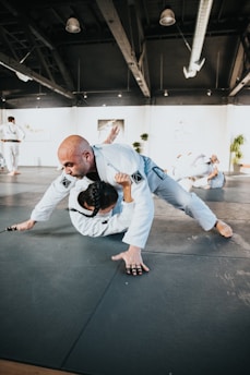 Two martial artists are practicing grappling techniques on a mat in a spacious training facility. The scene takes place under a high ceiling with exposed beams and bright lighting. Both individuals are wearing white martial arts uniforms, and the setting suggests a focus on physical technique and dynamic movement. Other people are visible in the background, engaged in similar training activities.