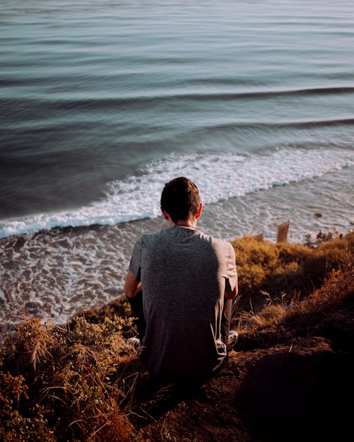 man in gray shirt sitting on brown grass near body of water during daytime
