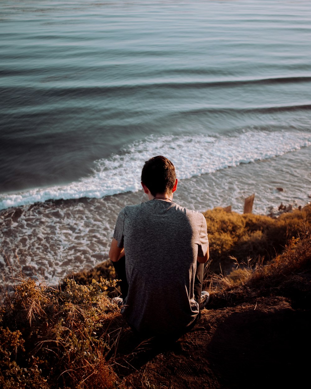 man in gray shirt sitting on brown grass near body of water during daytime