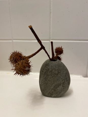 A minimalist stone vase with a smooth texture holds dry thorny branches, set against a tiled white wall.
