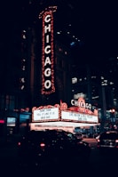 A nighttime scene featuring a large, illuminated theater sign with the word CHICAGO in bright lights. Below, a marquee advertises a sold-out performance by Dane Cook. The setting is urban, with cars on the street and the entrance to the theater in view.