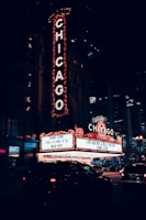 A nighttime scene featuring a large, illuminated theater sign with the word CHICAGO in bright lights. Below, a marquee advertises a sold-out performance by Dane Cook. The setting is urban, with cars on the street and the entrance to the theater in view.