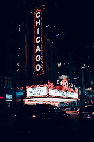 A nighttime scene featuring a large, illuminated theater sign with the word CHICAGO in bright lights. Below, a marquee advertises a sold-out performance by Dane Cook. The setting is urban, with cars on the street and the entrance to the theater in view.